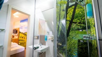 Un baño moderno con cabina de ducha de vidrio, un lavabo y una cama visible a través de una puerta abierta.