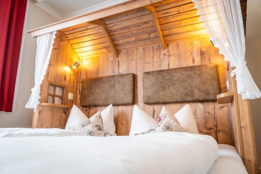 Una cama con ropa blanca y varias almohadas, un cabecero de madera con una lámpara, y una cortina roja a la izquierda.