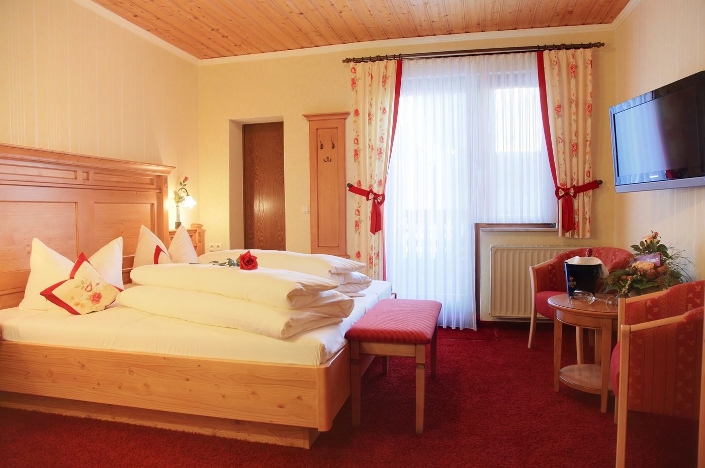 Habitación de hotel con una cama de madera, una mesita con una silla, un radiador y un televisor colgado en la pared.