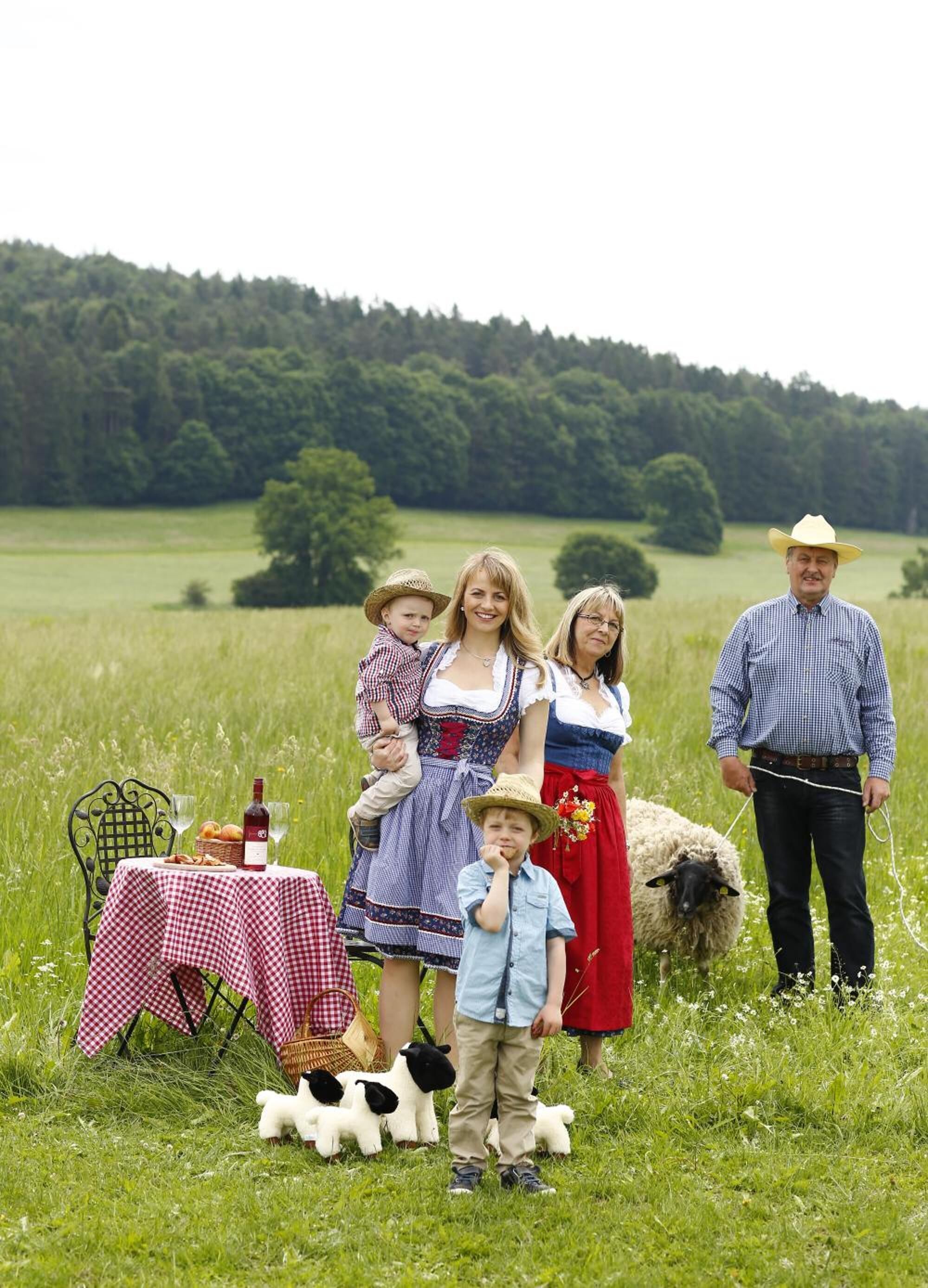 A family in a field with a picnic table, a bottle of wine, and a basket of bread.