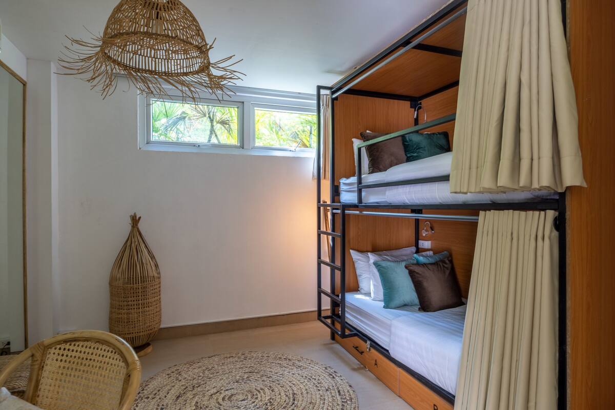 A bunk bed room with a woven ceiling light, a wicker chair, and a view of greenery outside.
