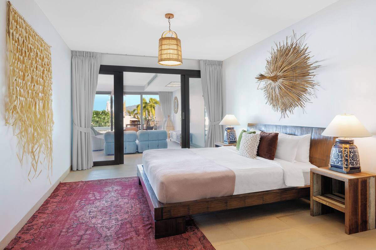 A bedroom with a large bed, two bedside tables with lamps, a decorative wall piece, and a view through an open door to an outdoor seating area.