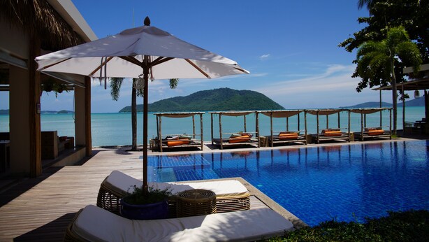 Zona junto a la piscina con sillones y sombrillas, con vistas a un mar azul cristalino y una isla distante.