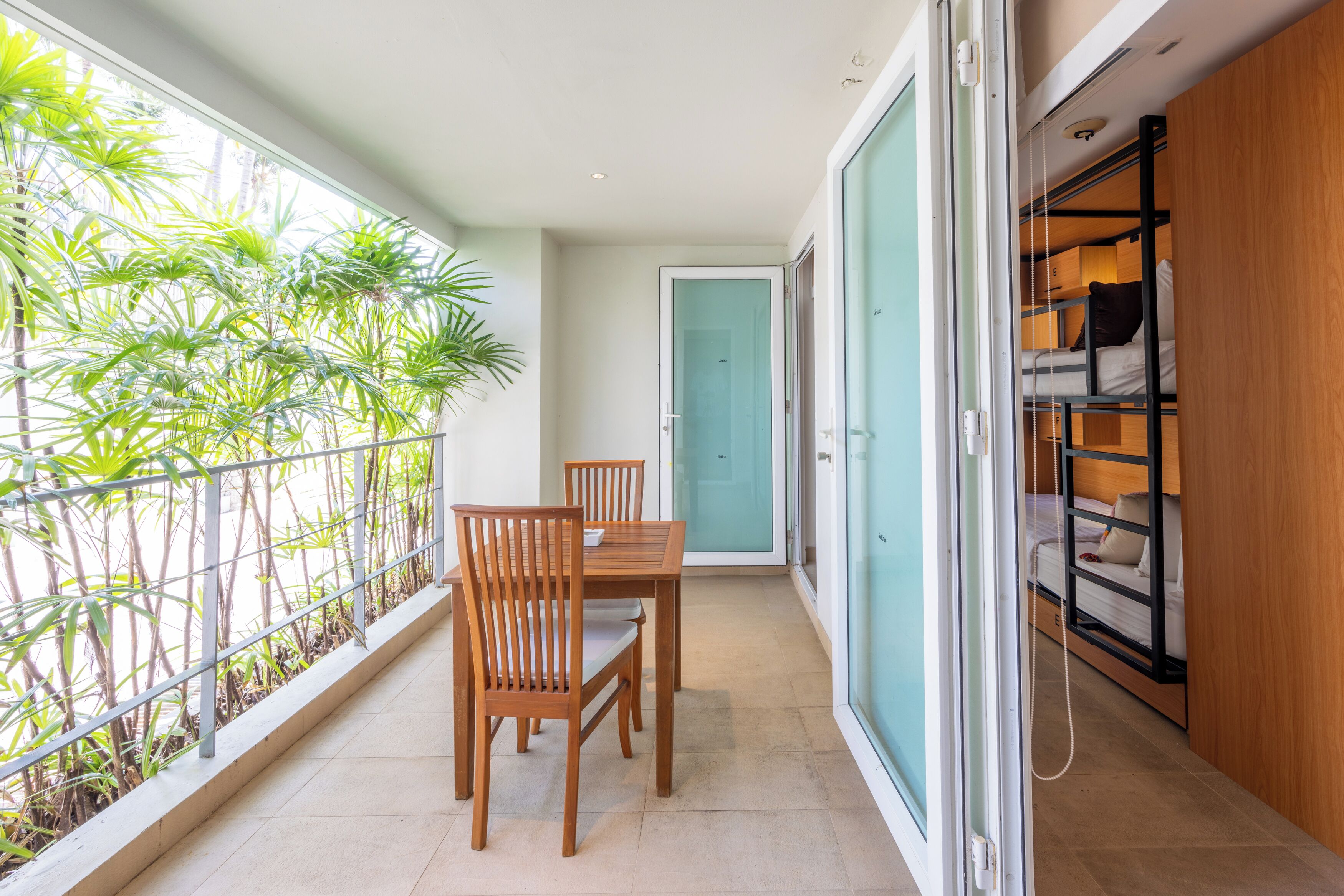A balcony with a dining table and chairs, a view of greenery, and a sliding door leading to a bedroom with bunk beds.