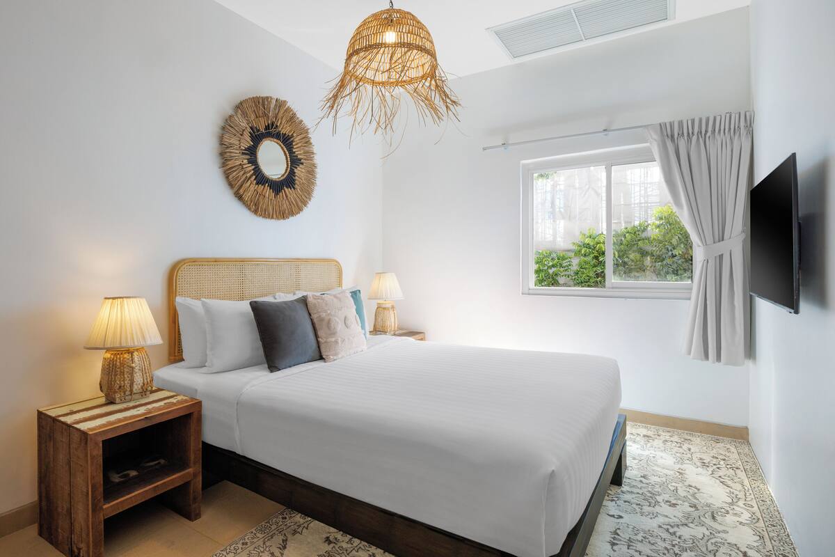 A bedroom with a bed, bedside lamps, a wall-mounted TV, a window with curtains, and a decorative mirror.