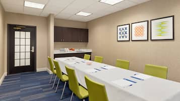 A conference room with a long table, green chairs, a white tablecloth, a sink, and framed artwork on the wall.