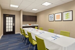 A conference room with a long table, green chairs, a white tablecloth, a sink, and framed artwork on the wall.