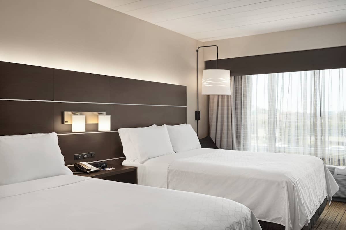 A hotel room with two beds, a bedside table with a phone, and a large window with curtains.