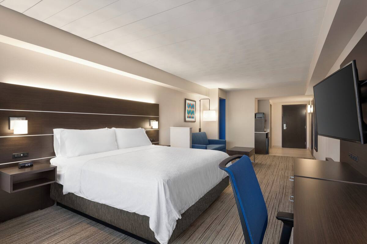 A modern hotel room with a large bed, bedside tables, a desk, a chair, a flat-screen TV, and a door.