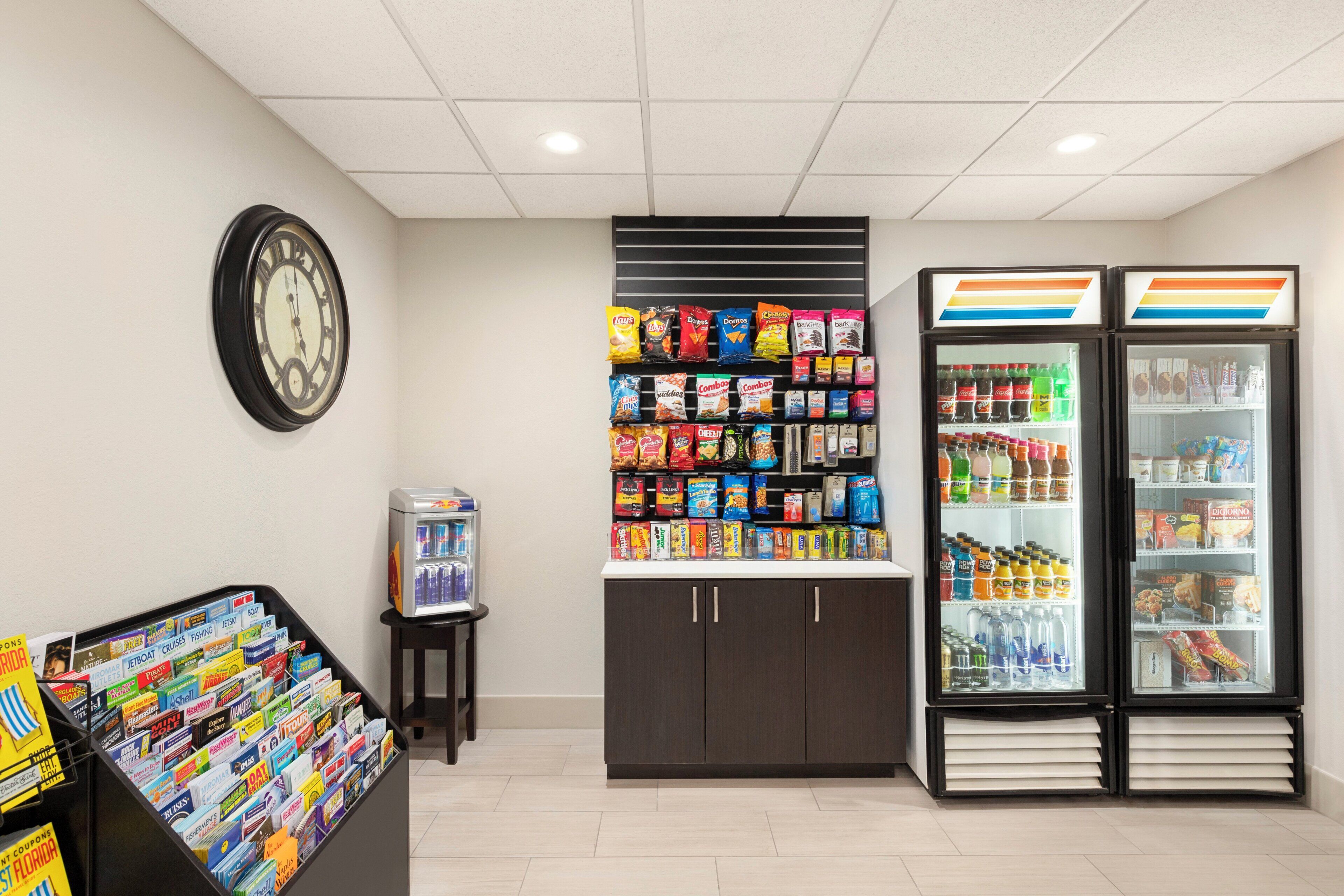A snack vending area with various snacks and beverages, a clock on the wall, and a display of travel brochures.