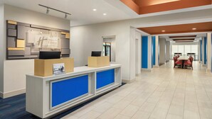 Un espace d’accueil d’hôtel au design moderne, comprenant un comptoir d’accueil, un écran numérique et un coin salon avec des tables et des chaises.