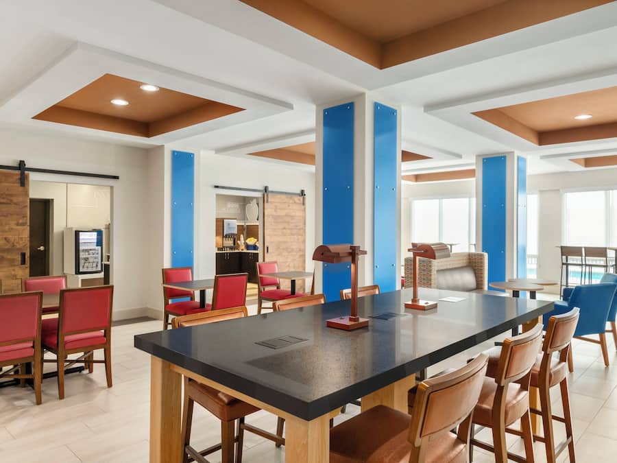 Un espacio interior amplio con una mesa larga y numerosas sillas, una zona de cocina al fondo y vistas al exterior.