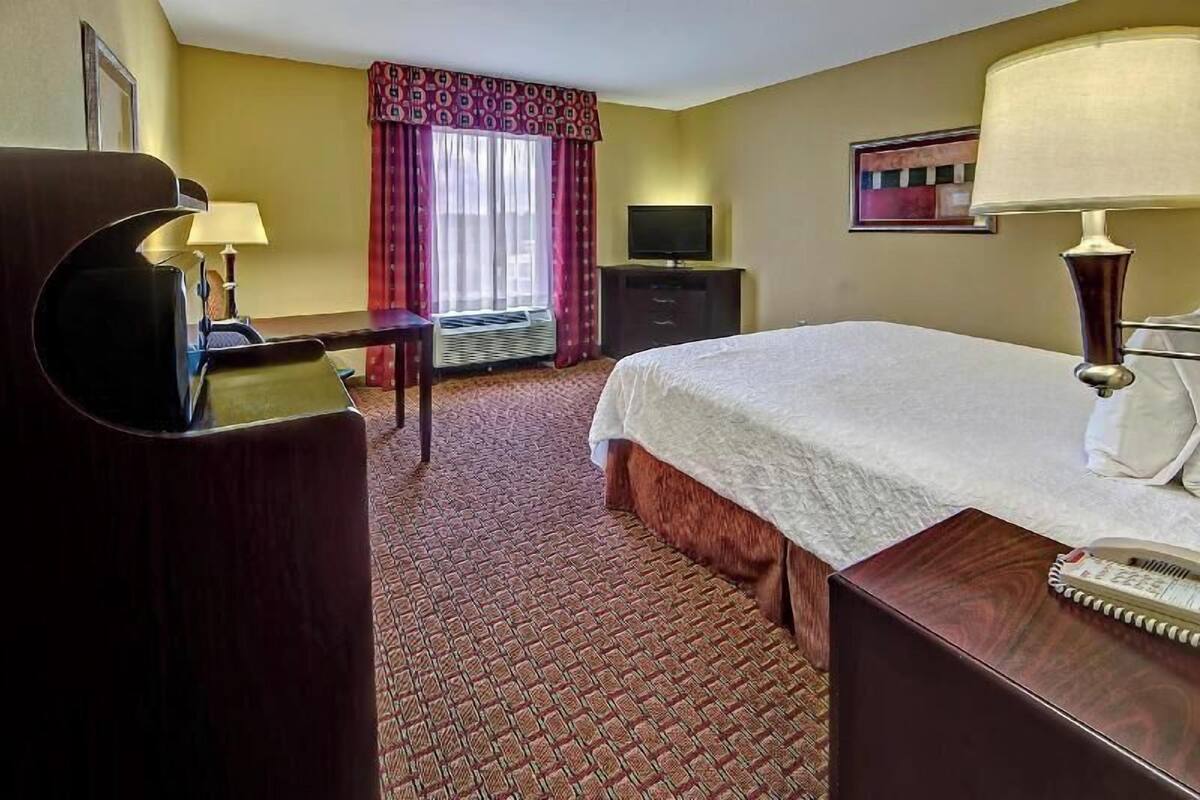 Una habitación de hotel con cama, escritorio, silla, televisor y lámpara.