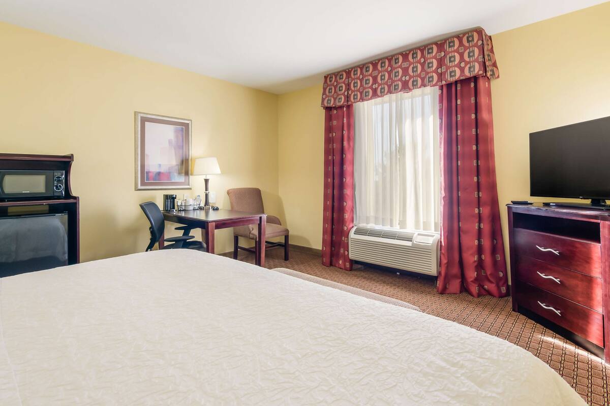 A hotel room with a large bed, a desk with a chair, a television, a microwave, and a window with curtains.