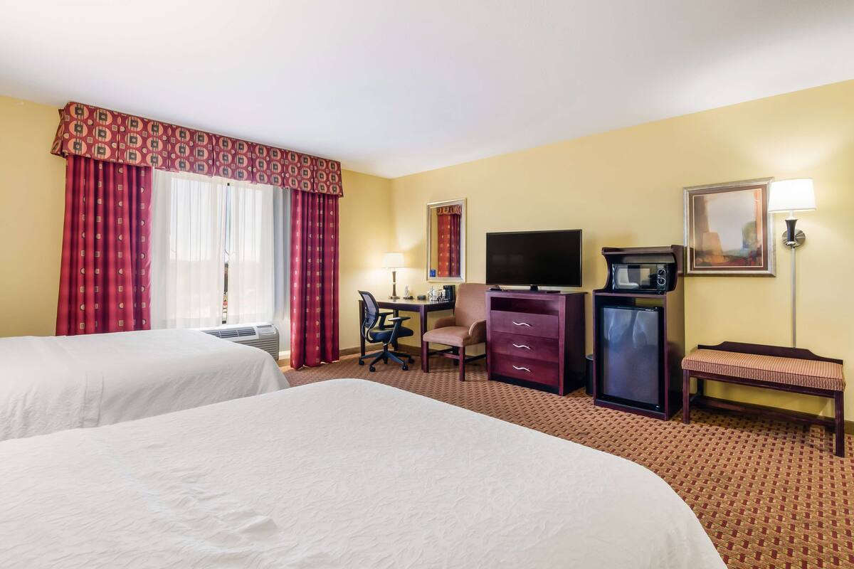 A hotel room with two beds, a desk with a chair, a television, a small refrigerator, and a bench.