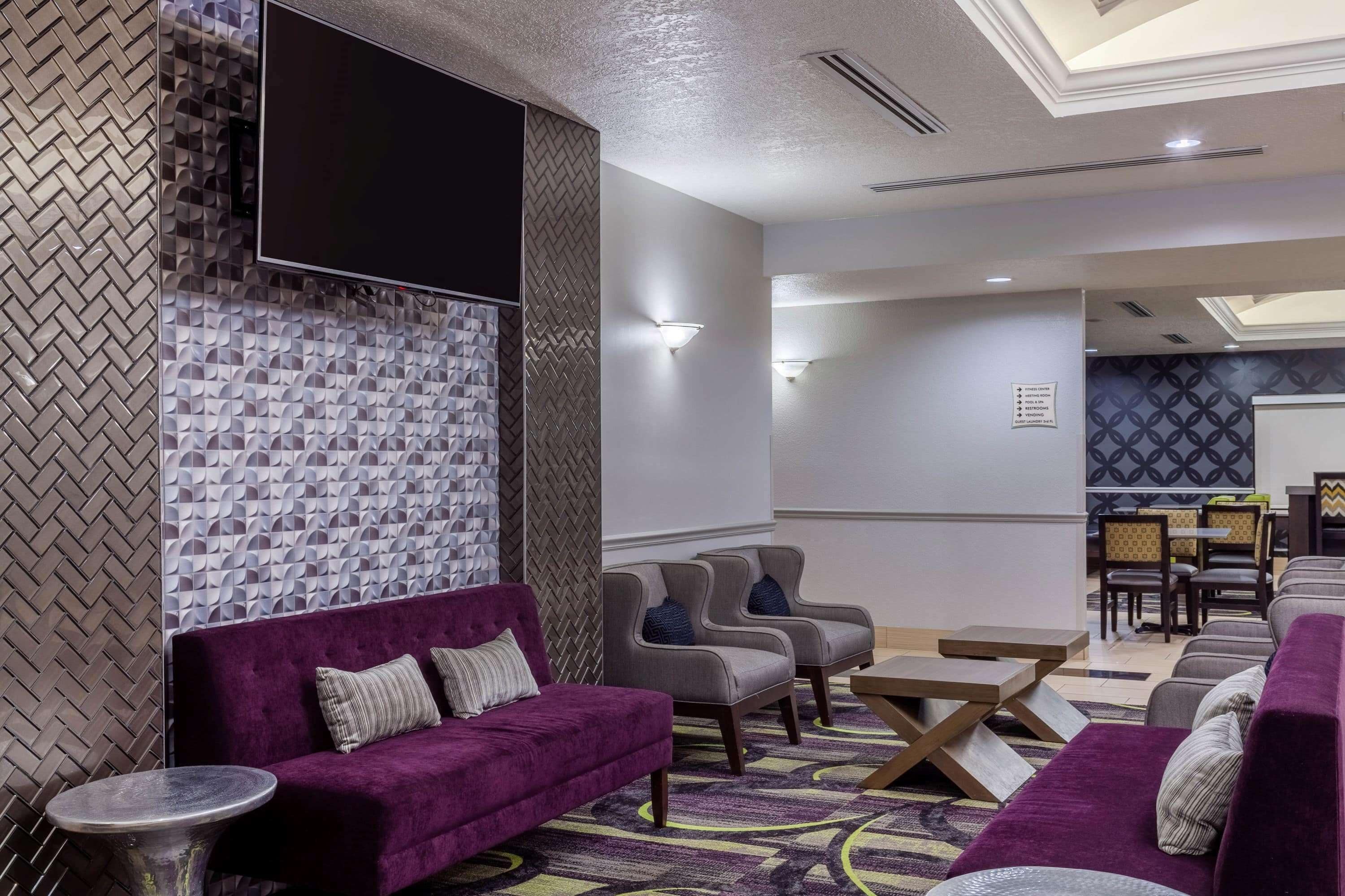 A modern hotel lobby with a seating area featuring purple sofas and grey armchairs, a flat-screen TV mounted on a textured wall, and a dining area with a patterned tablecloth.