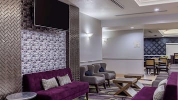 A modern hotel lobby with a seating area featuring purple sofas and grey armchairs, a flat-screen TV mounted on a textured wall, and a dining area with a patterned tablecloth.