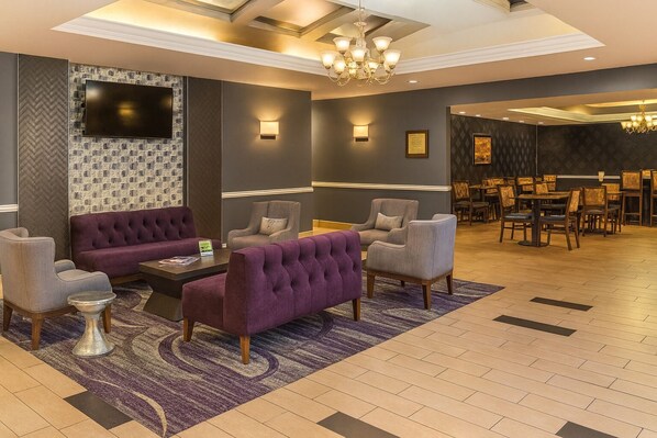A lobby with a seating area featuring a sofa, armchairs, and a coffee table. There is a television mounted on a textured wall, and a chandelier hangs from the ceiling.