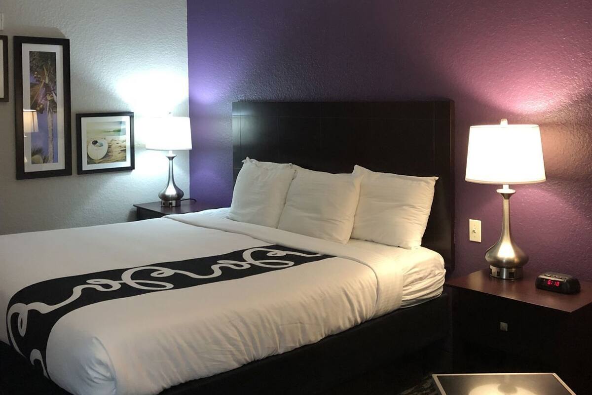 A hotel room with a bed, two bedside tables, a lamp, a clock, and framed pictures on the wall.