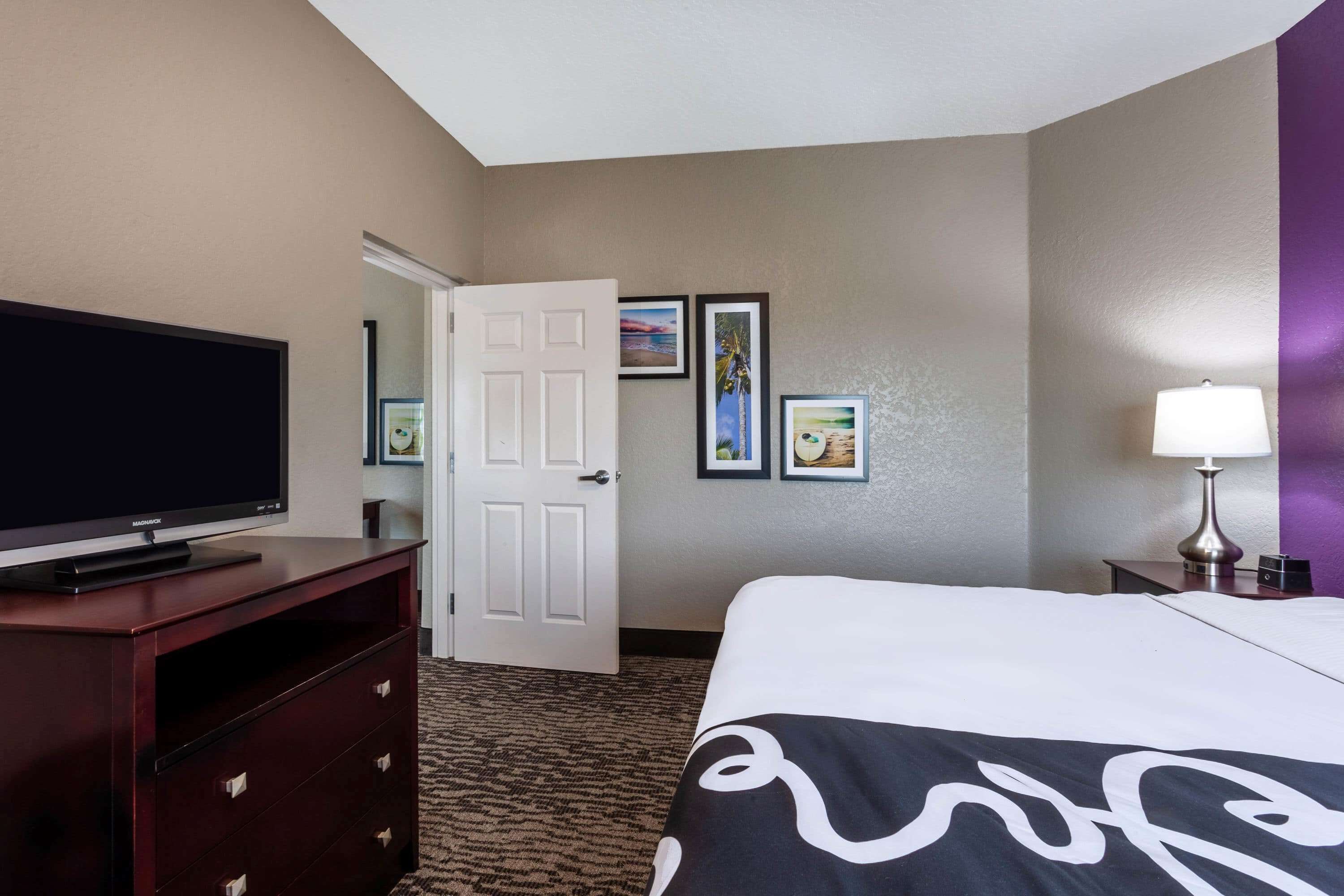A hotel room with a bed, a television on a wooden dresser, a door, and framed pictures on the wall.