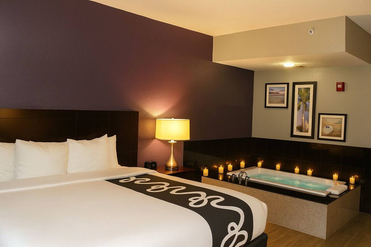 A hotel room with a large bed, a bedside table with a lamp, a bathtub with candles, and framed pictures on the wall.