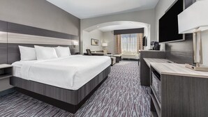 A modern hotel room with a large bed, a desk, a chair, a TV, and a bathroom visible through an arched doorway.