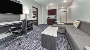 A modern hotel room with a flat-screen TV, a desk with a chair, a sofa, a small coffee table, and a kitchenette area.