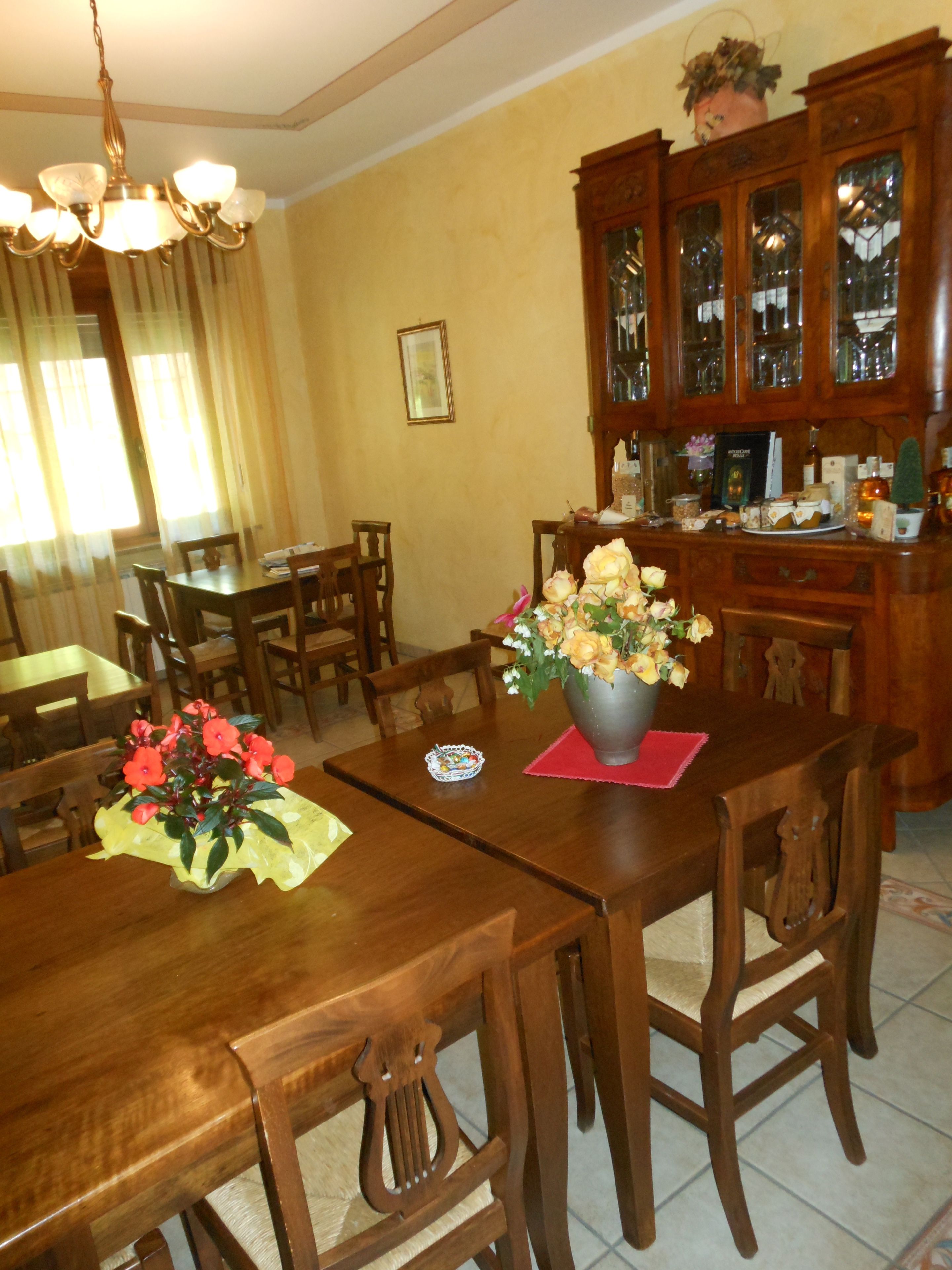 Un comedor con una mesa de madera, sillas y un jarrón de flores. Se observa una araña sobre la mesa y un aparador con puertas de cristal al fondo.