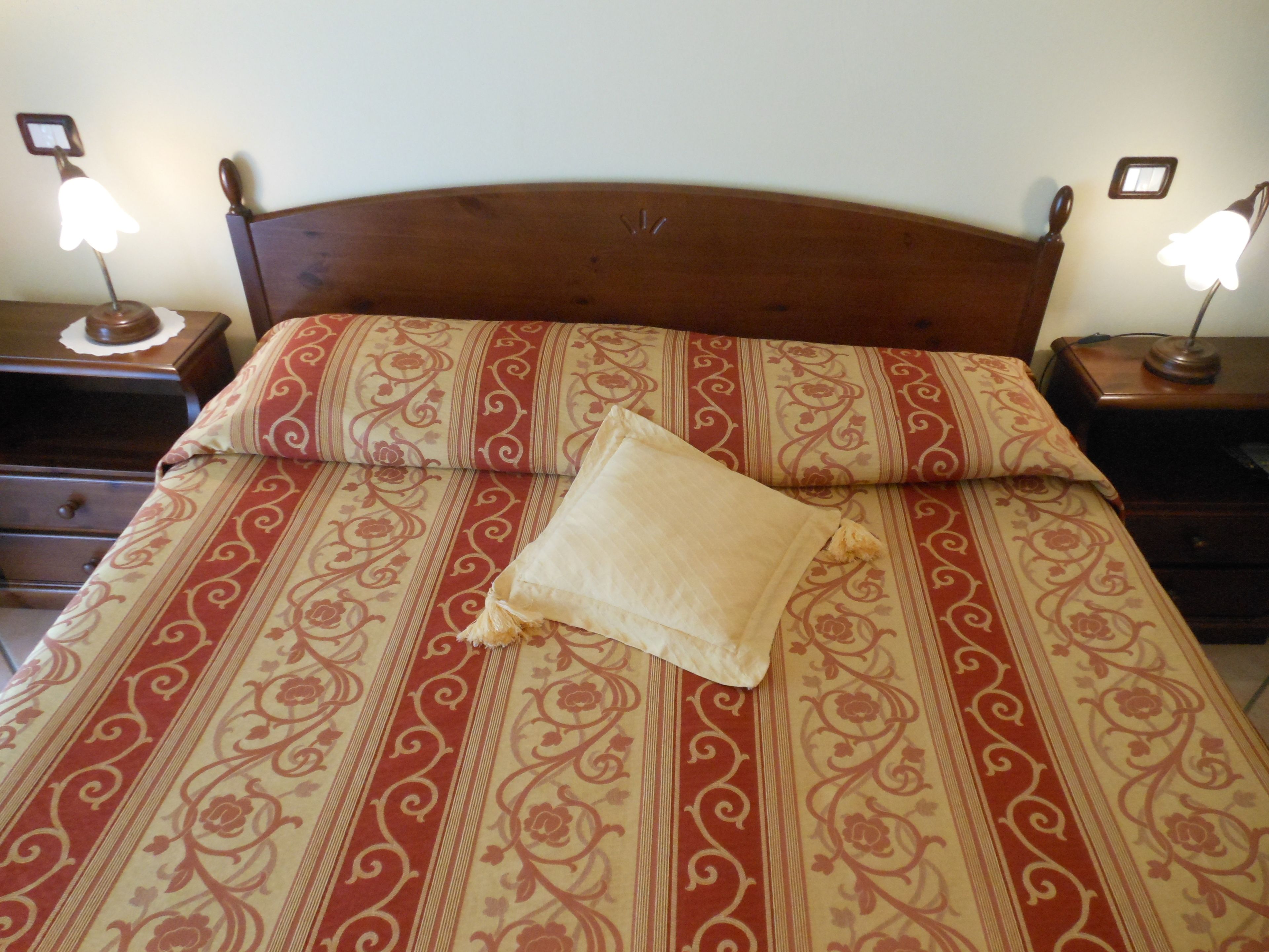 A neatly made bed with a patterned bedspread and a single pillow. There are two bedside tables with lamps on either side of the bed.