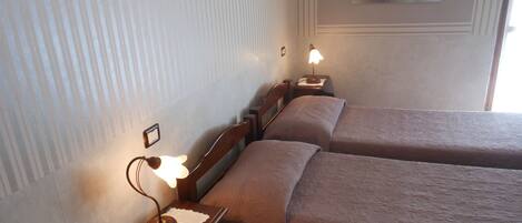 Habitación de hotel con dos camas individuales, una mesita de noche con lámpara, un cuadro enmarcado en la pared y una ventana con luz natural.