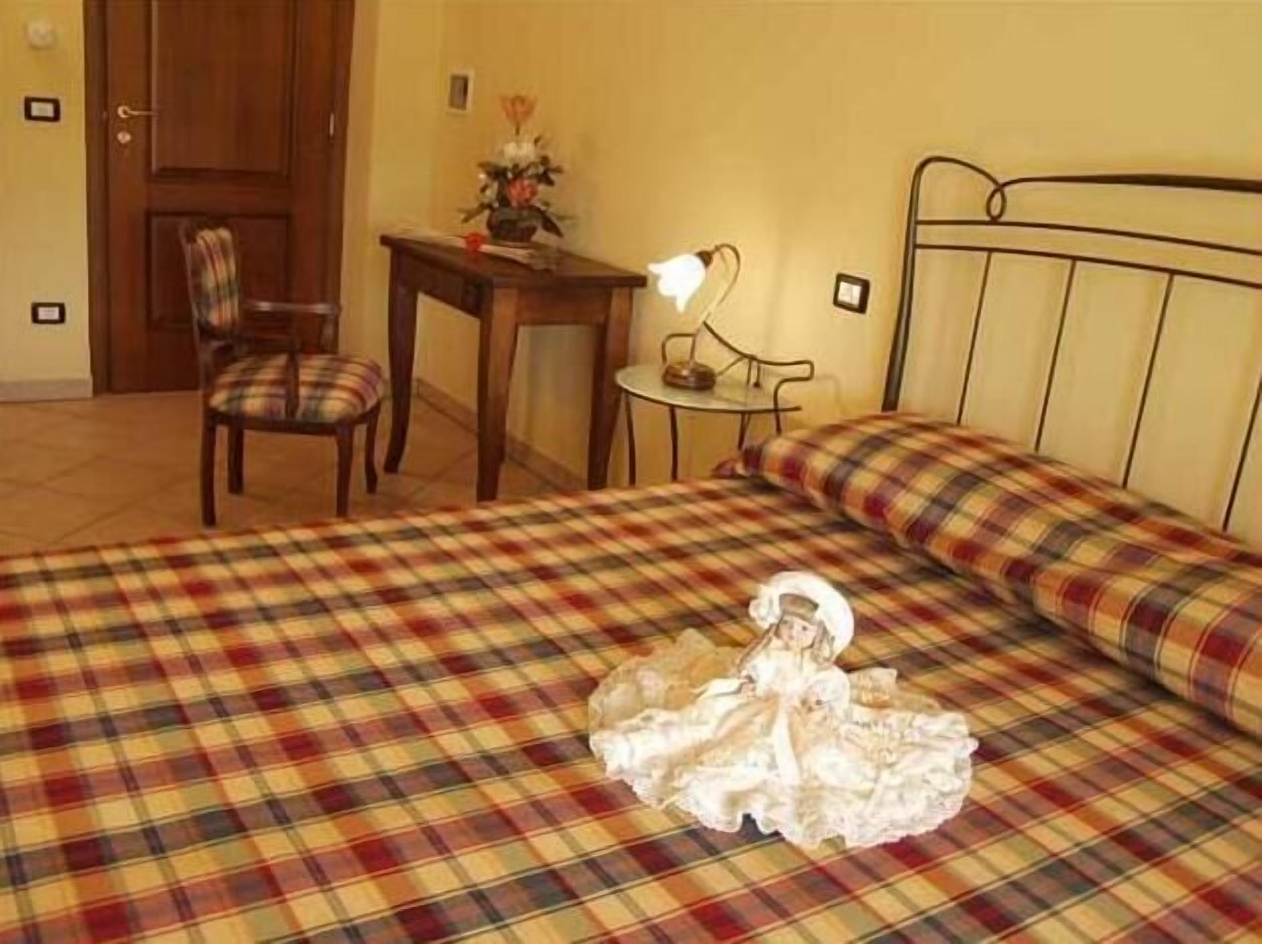 A room with a plaid bedspread, a small table with a lamp, a chair, and a decorative item on the wall.