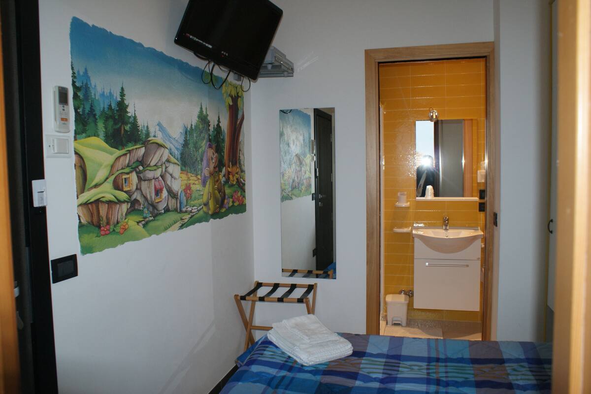 A bedroom with a bed, a painting, a television, a mirror, and a bathroom with a sink and toilet.