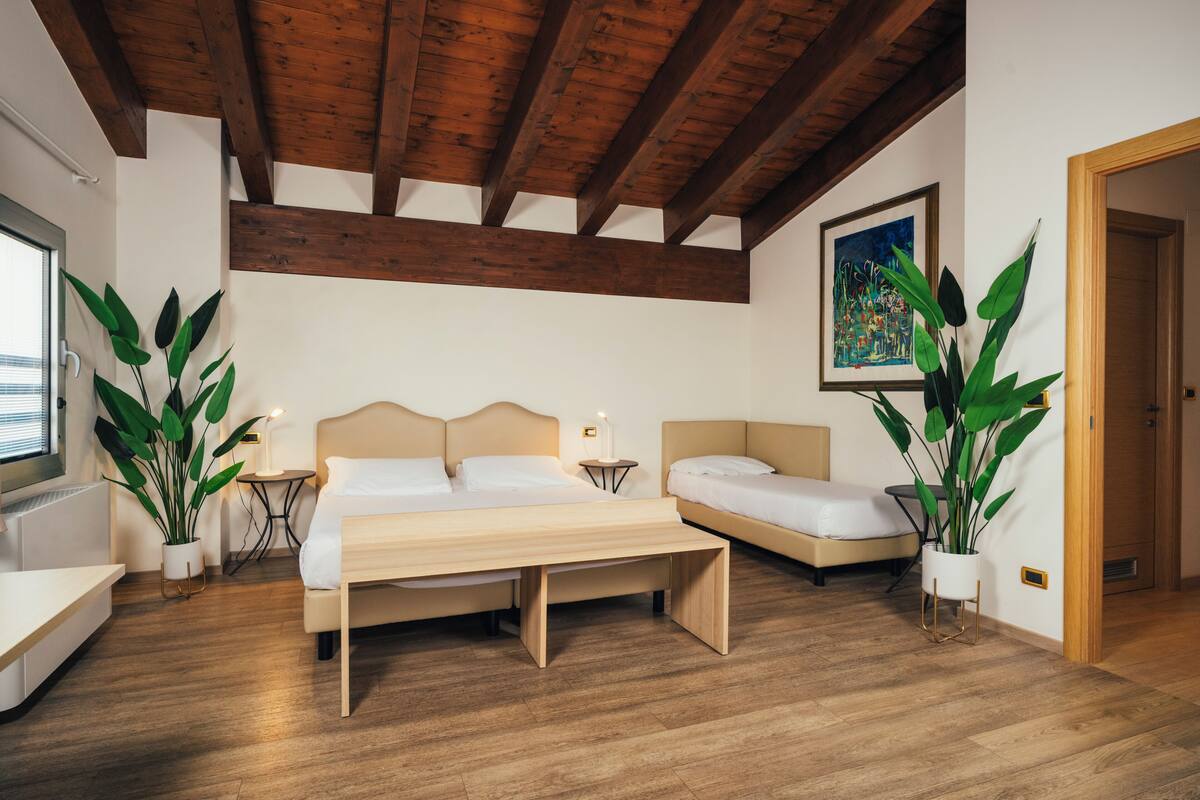A bedroom with a wooden ceiling, a bed, a bench, and two potted plants.