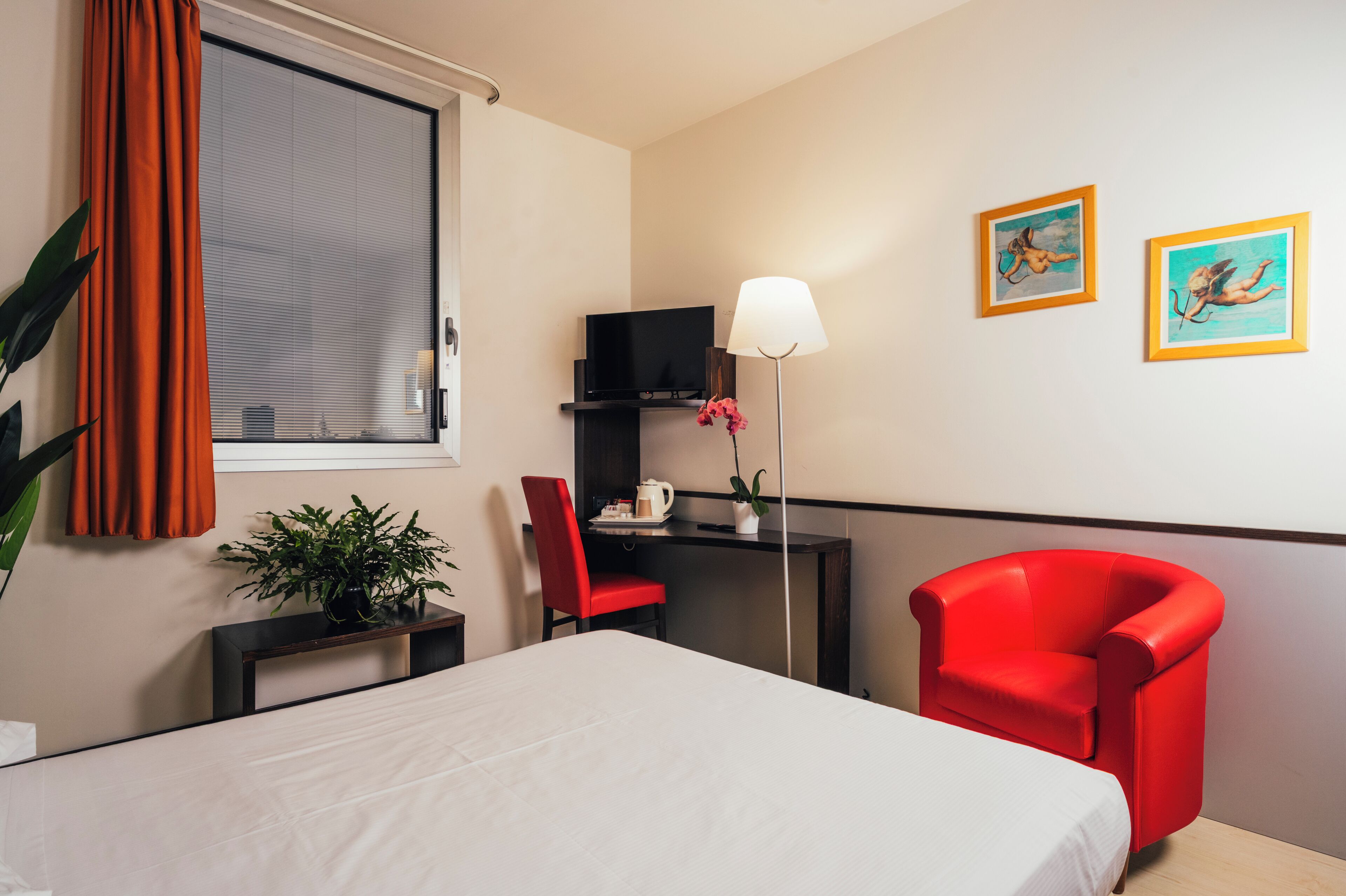 A hotel room with a bed, a red armchair, a desk with a lamp, a TV, and two framed pictures on the wall.