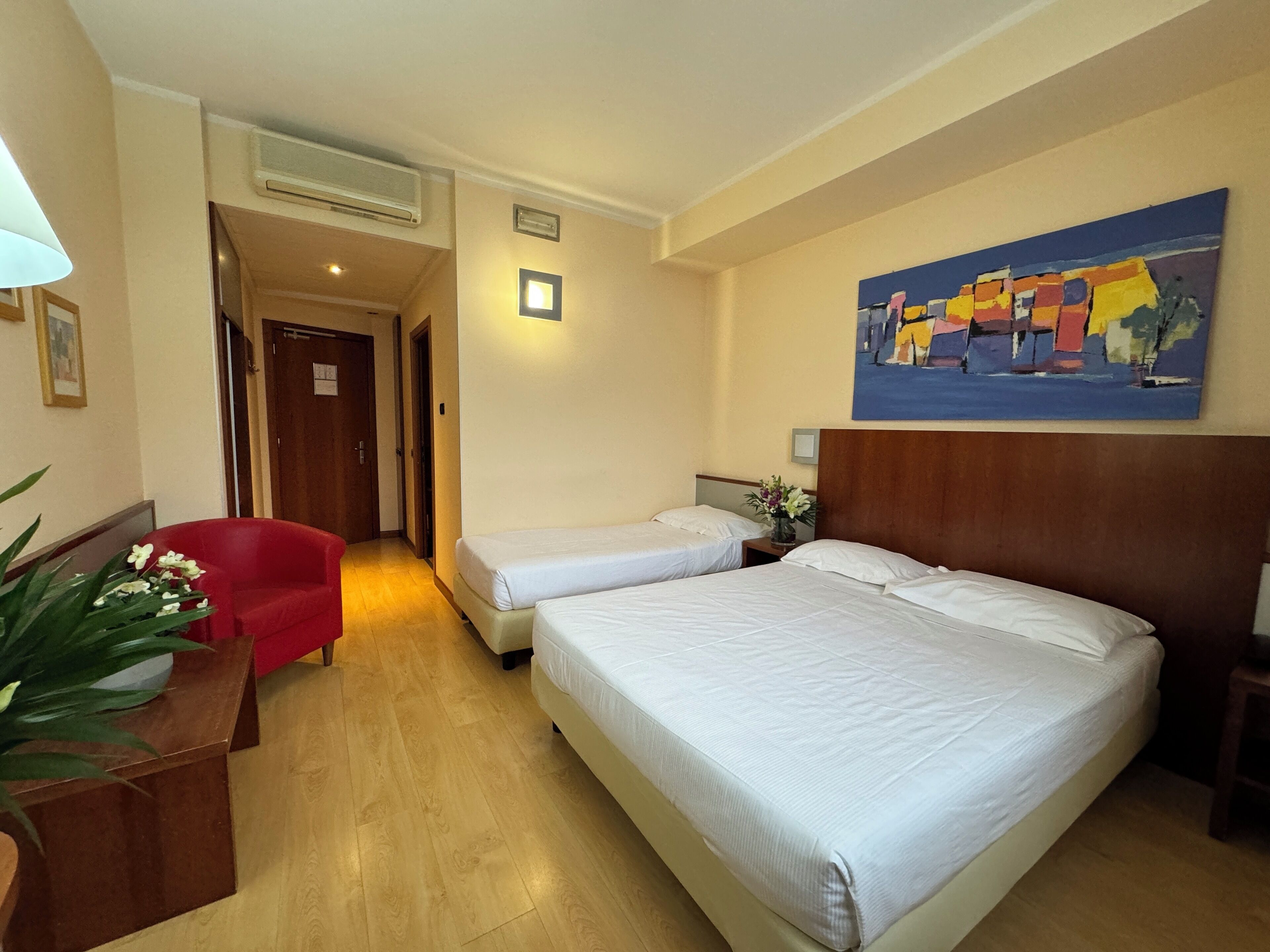 Ein Hotelzimmer mit zwei Betten, einem roten Sessel, einem Holfnachtkästchen, einem Bild an der Wand und einer Topfpflanze.