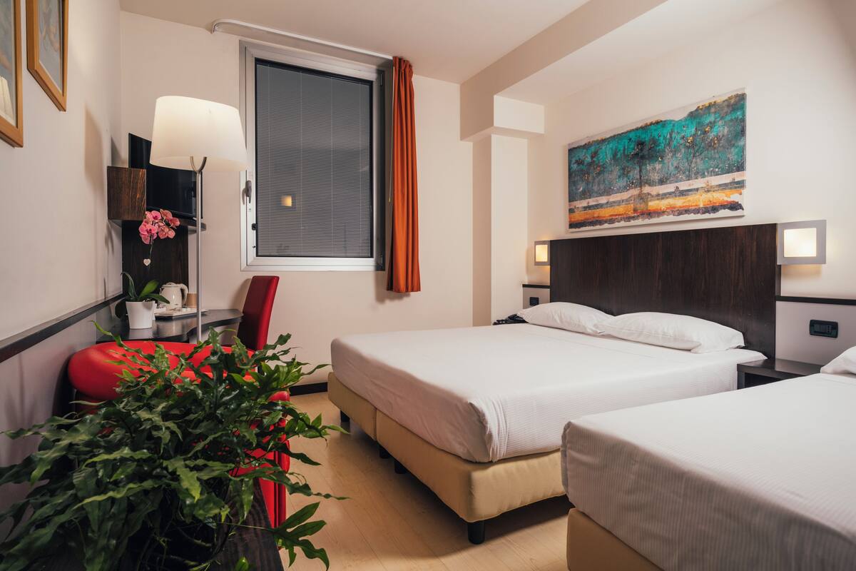 A hotel room with two beds, a large window with curtains, a painting on the wall, a red chair, a bedside table with a lamp, and a potted plant.