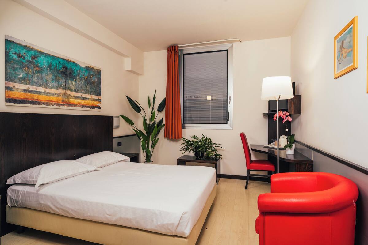 A hotel room with a bed, a desk, a chair, a window, and a painting on the wall.