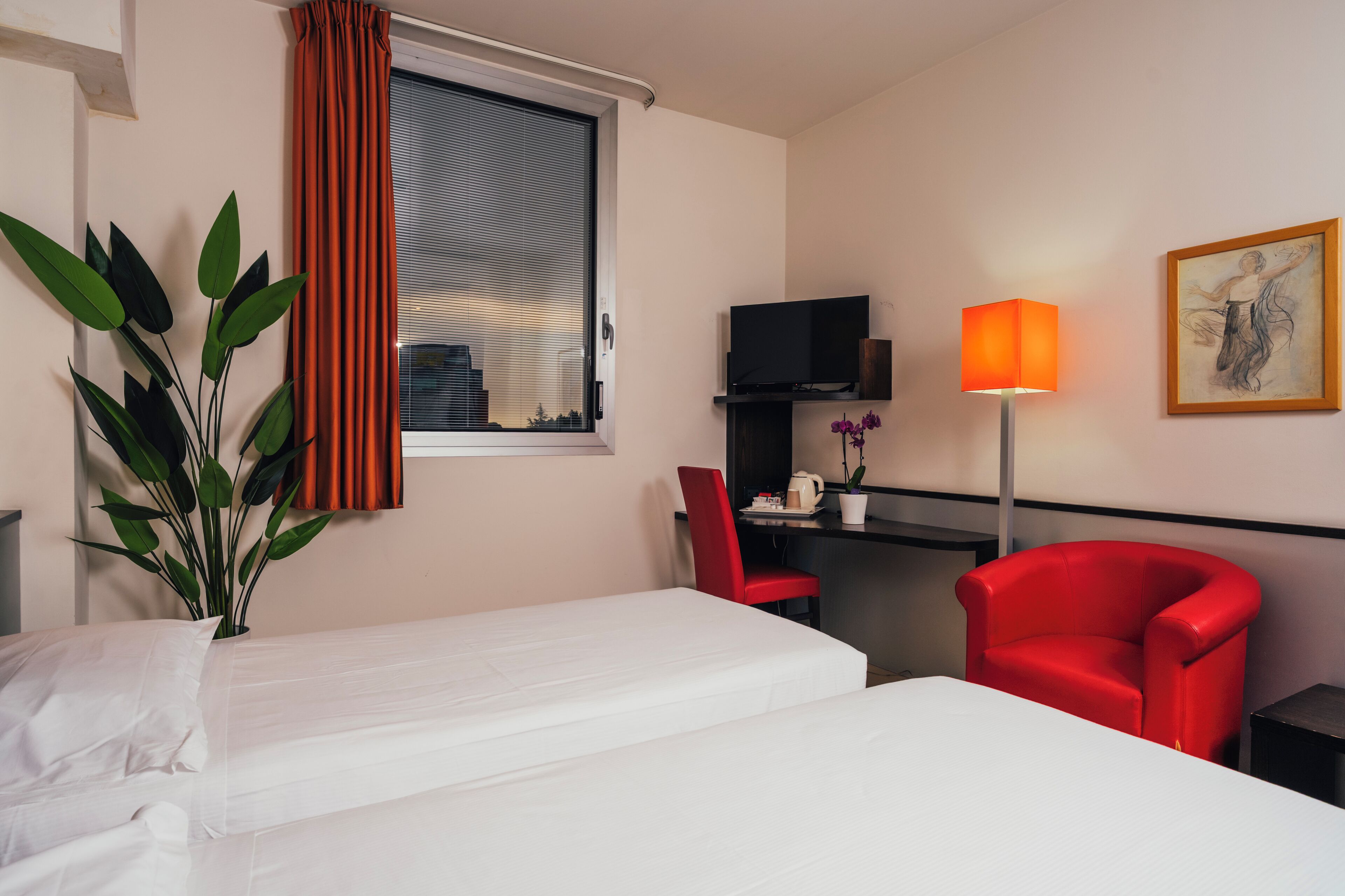 A hotel room with a bed, a desk with a chair, a TV, a lamp, a plant, and a painting on the wall.