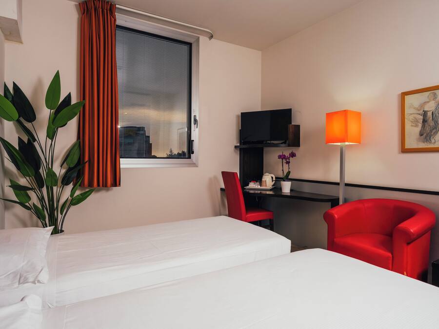 A hotel room with a bed, a desk with a chair, a TV, a lamp, a plant, and a painting on the wall.