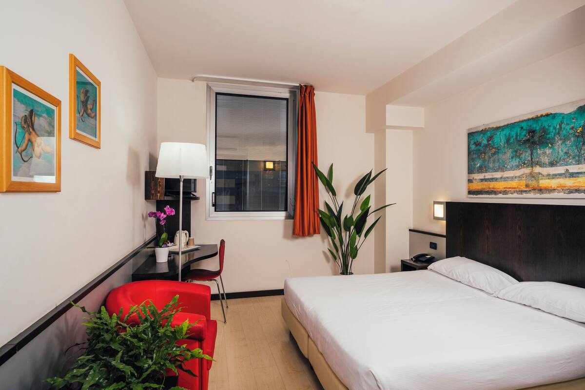 A hotel room with a bed, a desk, a chair, a lamp, a window with curtains, and a painting on the wall.