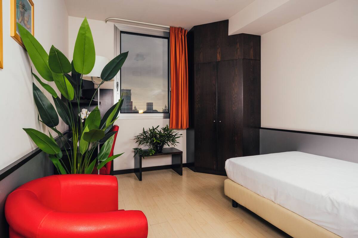 A compact hotel room with a bed, a red chair, a small table with a plant, and a window with curtains.