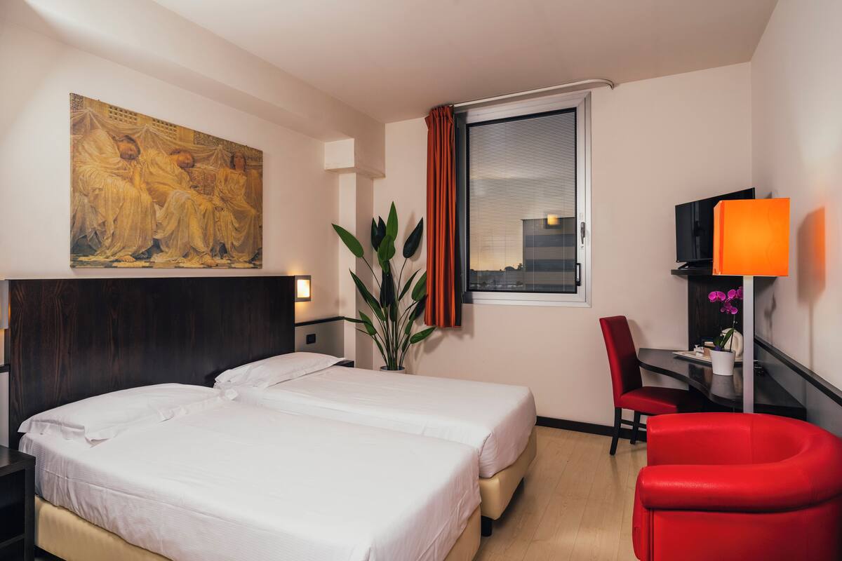 A hotel room with two beds, a desk with a chair, a TV, a plant, and a painting on the wall.
