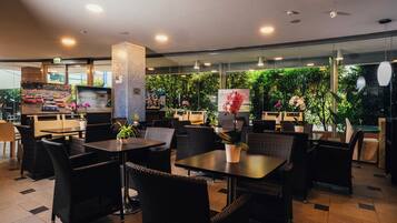 Um restaurante moderno com móveis de vime, plantas e uma grande janela com vista para o verde.