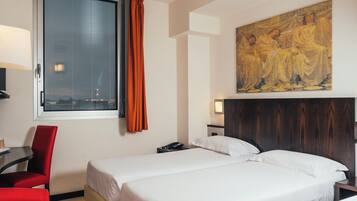 Quarto de hotel com duas camas, uma escrivaninha, uma cadeira, uma planta em vaso e um quadro na parede.