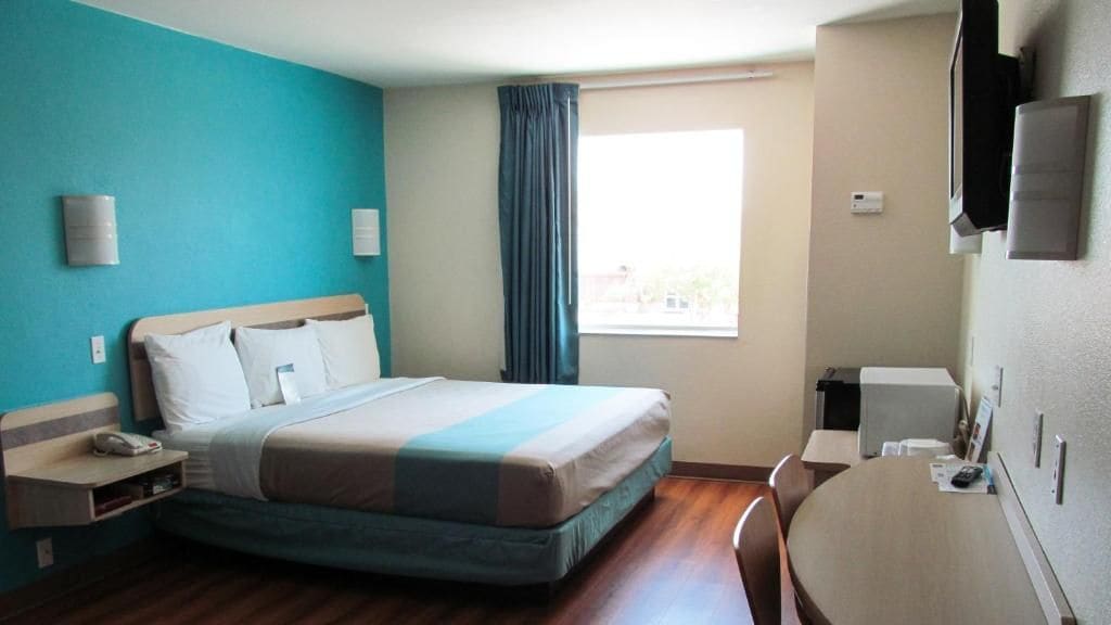 A hotel room with a bed, bedside table, chair, desk, and a window with curtains.