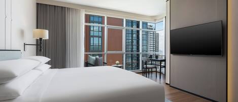 A hotel room with a large bed, a flat-screen TV, a desk with a chair, and a view of the city through large windows.