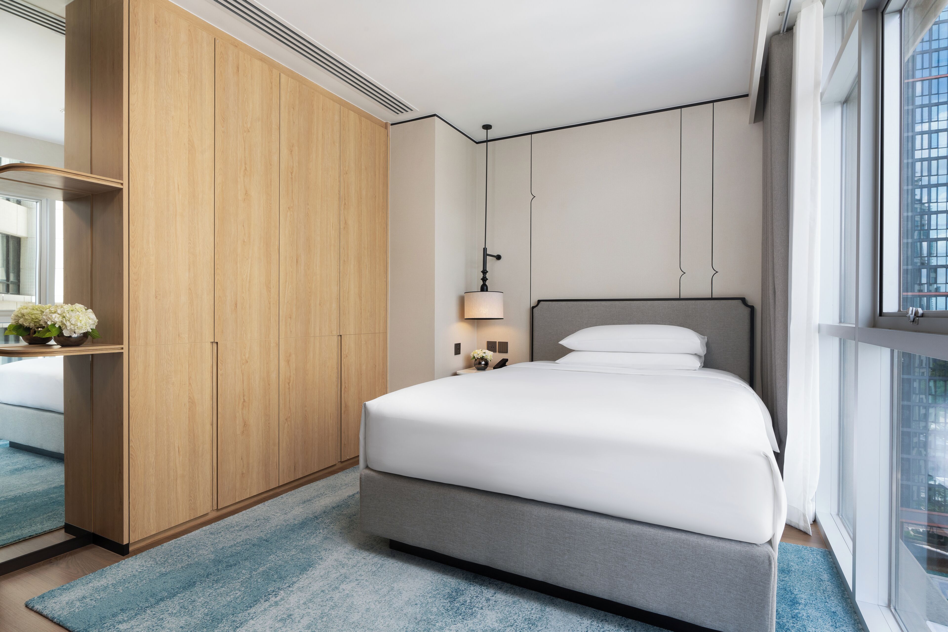 Un dormitorio moderno con una cama grande, un armario, una mesita de noche con lámpara y vistas a la ciudad desde la ventana.