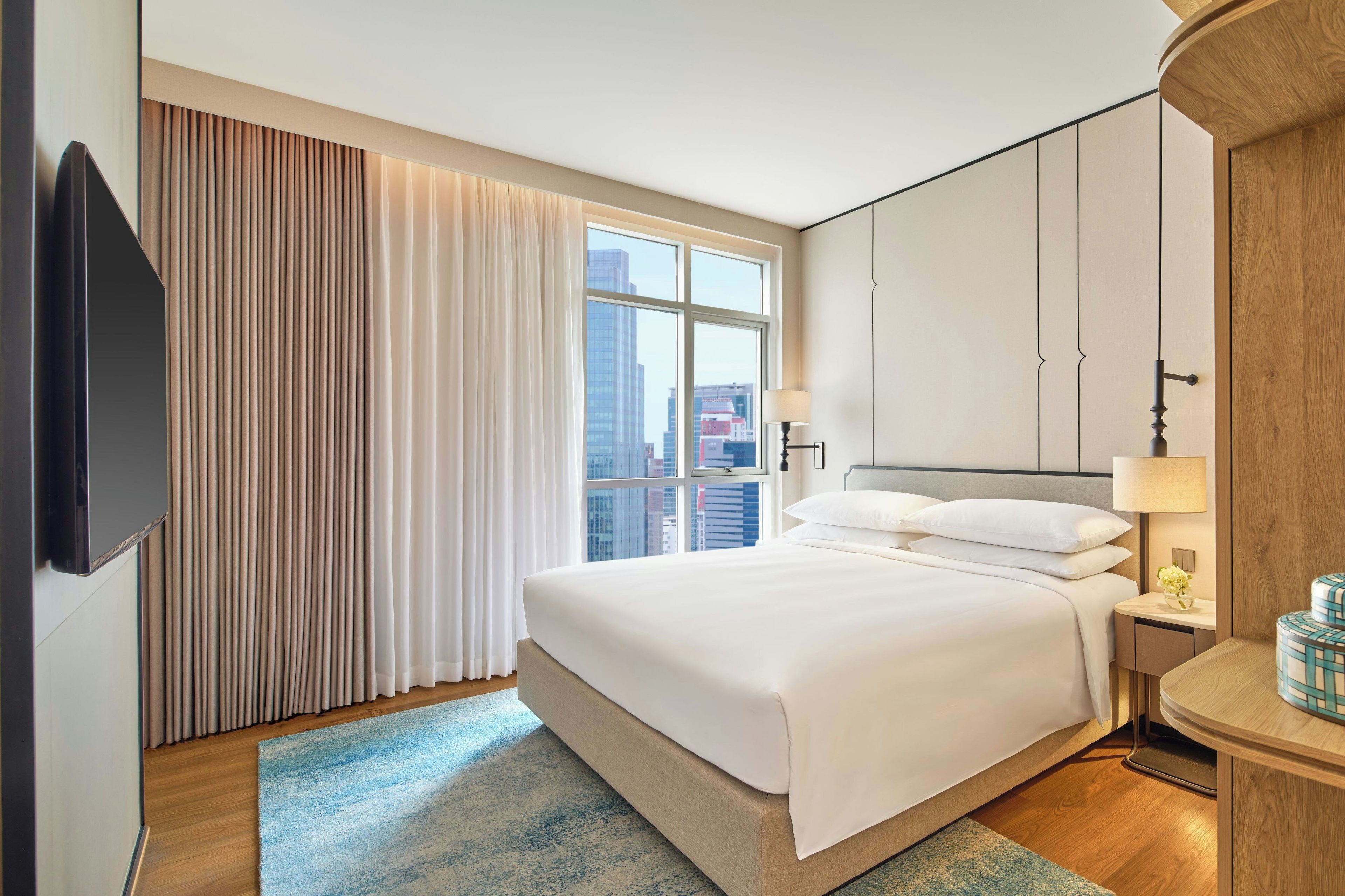 Une chambre d’hôtel avec un grand lit, un téléviseur à écran plat fixé au mur, une fenêtre avec des rideaux et une vue sur le paysage urbain.
