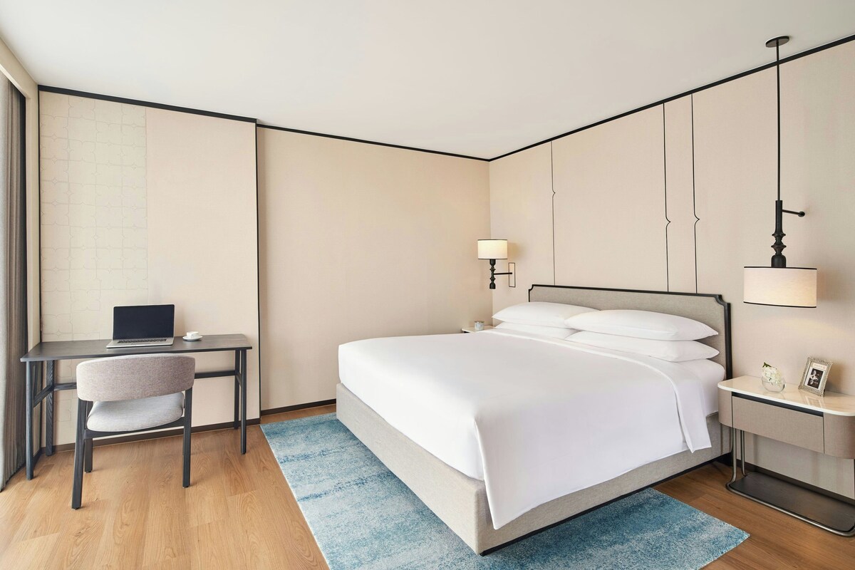 A hotel room with a bed, a desk, a chair, a nightstand, and a lamp.