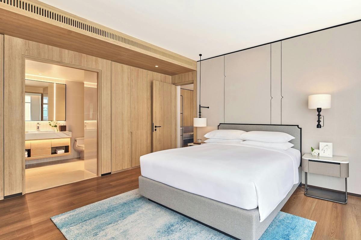 A modern hotel room with a large bed, a bedside table with a lamp, a nightstand, a mirror, and a wooden wardrobe.